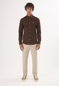 Camicione marrone a maniche lunghe con colletto button-down, bottoni bianchi e logo ricamato; abbinato a pantaloni beige e sneakers di colore chiaro.
