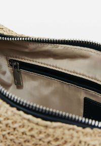 Das Innere der geflochtenen Strohtasche ist mit einem beigen Satinfutter ausgestattet und verfügt über eine kleine Reißverschlusstasche sowie silberne Reißverschlussbeschläge entlang der oberen Kante.