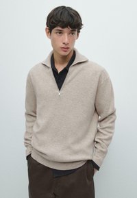 Maglione beige lavorato a maglia con colletto a mezza zip, texture a coste e maniche lunghe, indossato sopra una camicia scura con colletto, abbinato a pantaloni marroni.