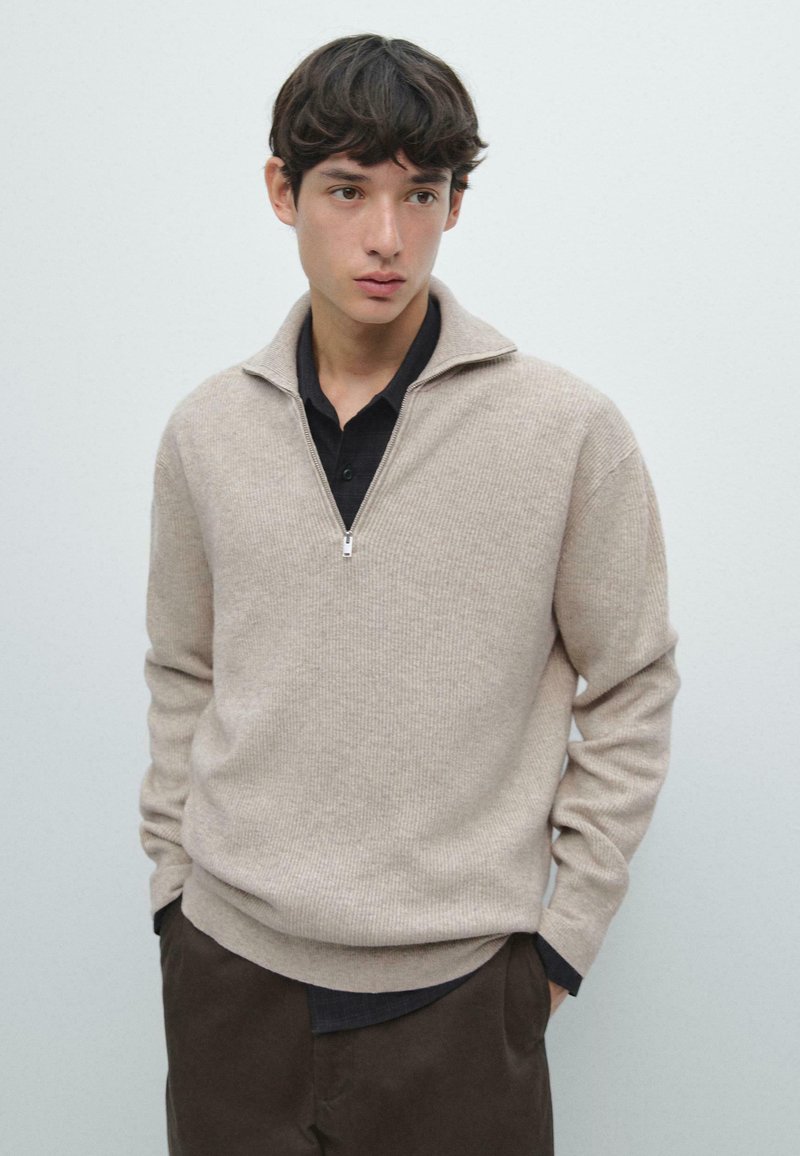 Maglione beige lavorato a maglia con colletto a mezza zip, texture a coste e maniche lunghe, indossato sopra una camicia scura con colletto, abbinato a pantaloni marroni.