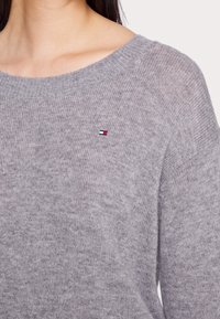Maglione grigio a maglia con un ampio scollo. Presenta una leggera texture e un piccolo logo ricamato in rosso, bianco e blu sul lato sinistro del petto.