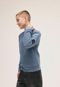 Niebieski sweter o fakturze z okrągłym dekoltem i długimi rękawami, z czarną metką na lewym ramieniu. Połączony z ciemnymi dżinsami.