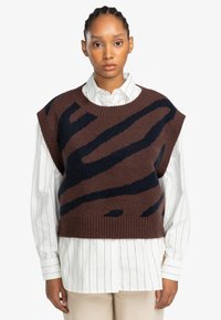 Braun gestrickter ärmelloser Pullover mit marineblauen abstrakten Streifen, einem gerippten Halsausschnitt, getragen über einem weißen karierten Hemd. Weiche Textur.