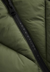 Olivgrön puffjacka med diamantquiltmönster, som har en svart dragkedja och präglad logotyp. Slät, matt textur.