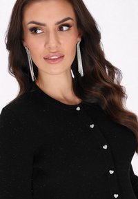 Musta ribbed-neulecardigan, jossa on sydämenmuotoiset hopeanväriset napit ja teksturoitu neulekuvio. Pitkät hiukset ja säihkysosiaat täydentävät tyyliä.