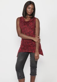 Top sans manches rouge avec un motif de feuilles et un col drapé, associé à un jean foncé avec des broderies décoratives sur les poignets.