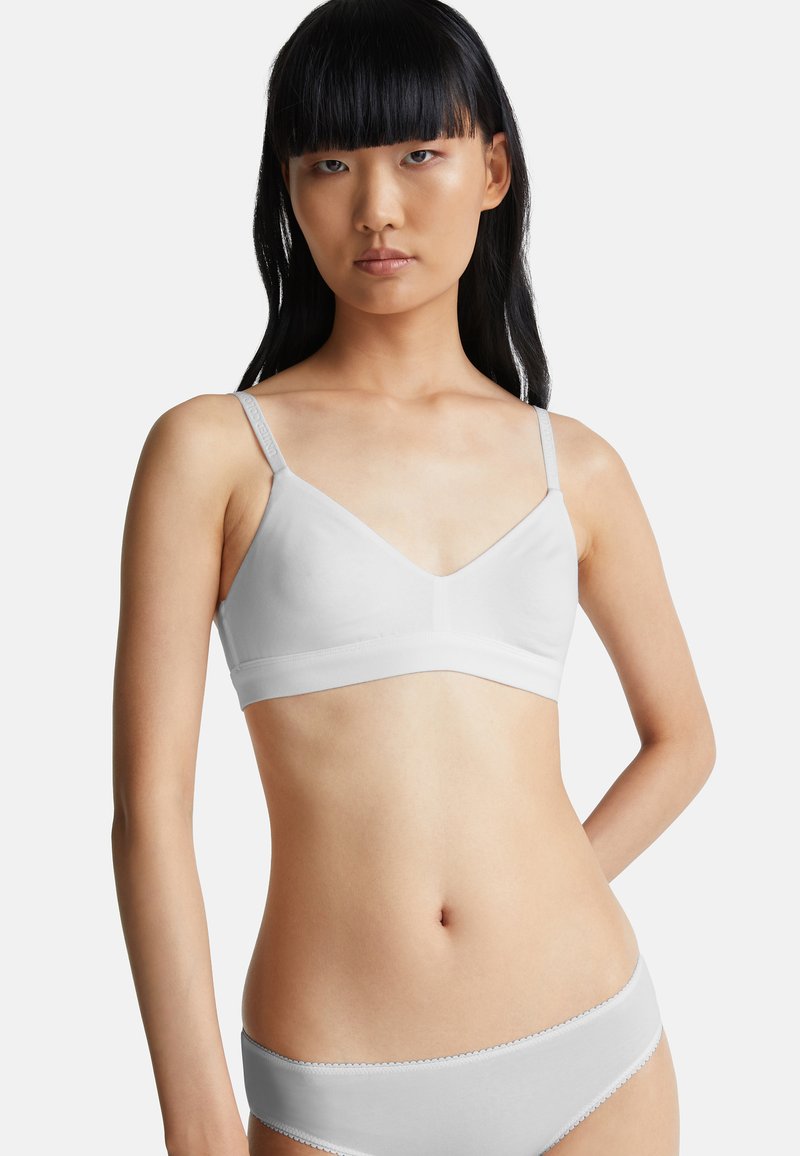 Mladá žena s dlouhými černými vlasy, oblečená v bílé bezdrátové podprsence a odpovídajícím spodním prádle, stojící proti jednoduchému světlému pozadí.