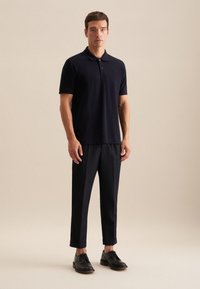 Granatowa koszulka polo z klasycznym kołnierzykiem, w zestawieniu z dopasowanymi czarnymi spodniami i ciemnymi skórzanymi butami, stojąca na neutralnym tle.