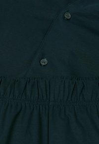 Donkergroene stof met een zachte textuur, voorzien van twee knopen en eenGathered elastische tailleband. Het ontwerp is eenvoudig en utilitair.