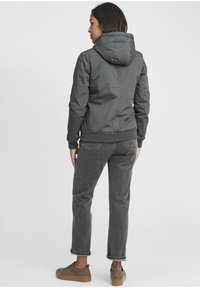 Gray hooded jacket with a smooth, water-resistant fabric, elastic cuffs, and a fitted silhouette, paired with dark denim jeans and brown shoes.