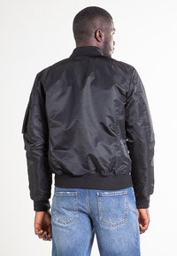 Czarna kurtka typu bomber z nylonu z prążkowanymi mankietami i ściągaczem u dołu, z boczną kieszenią. Nosi się ją z niebieskimi dżinsami, prezentując gładką fakturę.