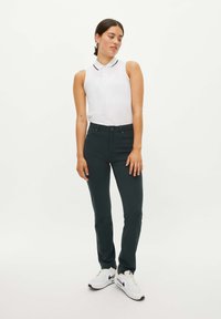 Pantaloni slim-fit verde scuro abbinati a un polo senza maniche bianca con colletto nero e dettagli sui bottoni, completati da sneaker bianche.