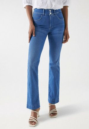 Personne portant un jean bleu taille haute avec fermeture à double bouton, associé à des sandales blanches à talons et brides, debout devant un fond uni.