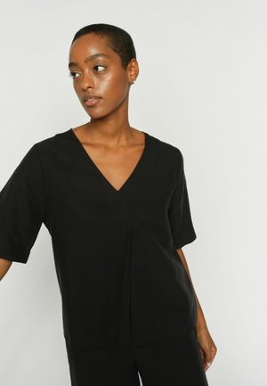 Kobieta o krótkich włosach, ubrana w czarną bluzkę z krótkim rękawem i dekoltem w serek, patrząca w lewo na tle jednolitego jasnego tła.