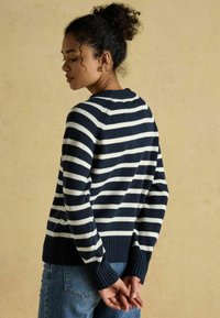 Kortsuksel juustega naine, kellel on bun soeng, kannab tumesinist ja valget triibulist kampsunit ning teksaseid, seistes beige seina vastas, käed selja taga kokku seotud.