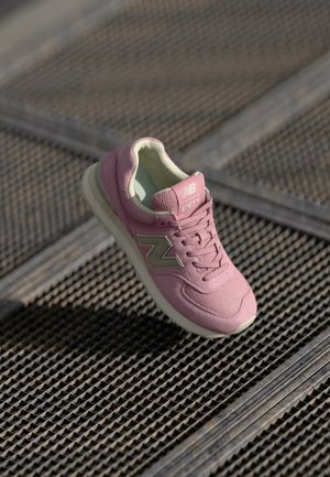 Różowy but sportowy New Balance 574 z beżowym logo na teksturowanej metalowej powierzchni, oświetlony naturalnym światłem.