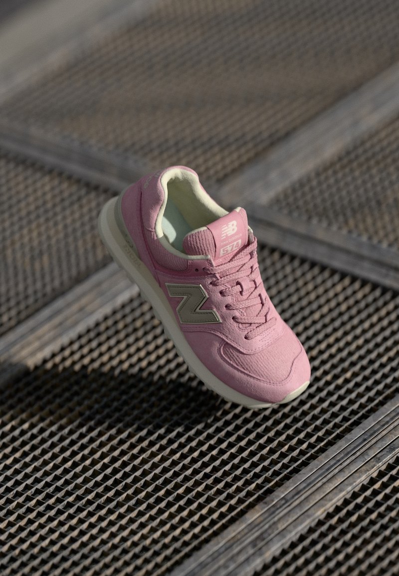 Baskets New Balance 574 roses avec logo beige sur une surface métallique texturée, éclairées par la lumière naturelle.