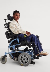 Fauteuil roulant électrique avec un châssis bleu, un siège noir, des roues argentées et des poignées texturées. L'utilisateur porte une chemise rayée et des chaussures beiges.