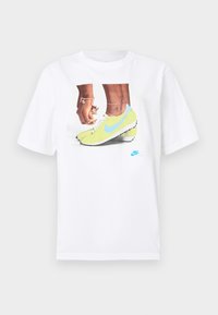 Balts kokvilnas T-krekls ar grafiku, kurā attēloti zili akcentēti dzelteni Nike zābaki un kreisajā rokā valkāta rokassprādze.