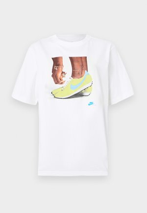 Hvit bomull t-skjorte med et grafisk motiv av føtter i gule Nike fotballsko med blå detaljer, kombinert med et armbånd på venstre håndledd.