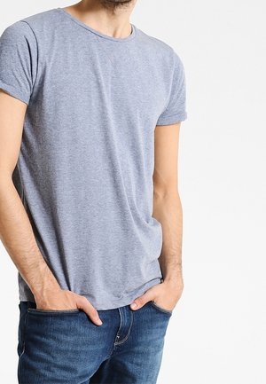 Mężczyzna w jasnoszarej koszulce z krótkim rękawem i niebieskich dżinsach, z rękami w kieszeniach na jednolitym białym tle.