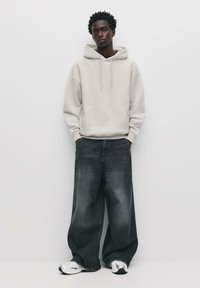 Felpa grigia chiara con tasca frontale, abbinata a pantaloni in denim scuro larghi. Il modello indossa scarpe sportive argento e si trova contro uno sfondo bianco.