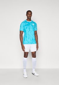 Τιρκουάζ ποδοσφαιρική φανέλα με λευκές λεπτομέρειες, που διαθέτει υφή σχεδίασης. Συνδυάζεται με λευκά σορτς και κάλτσες μέχρι το γόνατο, καθώς και με ποδοσφαιρικά παπούτσια.