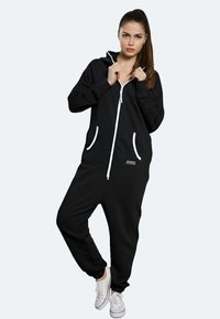 Tuta nera con zip realizzata in morbido tessuto, con accenti bianchi, tasche laterali e una vestibilità rilassata. Abbinata a sneakers bianche.