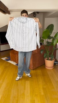 Pásikava košulja z gumbi v beli barvi s temnimi črtami; ima ovratnik, dolge rokave in ravno kroj. Model jo drži ob ozadju.