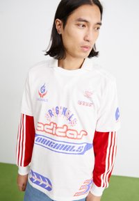 Balta Adidas Originals marškinėliai su raudonais ir mėlynais akcentais, su dideliu grafiniu tekstu ir juostelėmis ant rankovių. Pagaminti iš medvilnės, laisvas kirpimas.