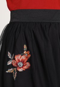 Jupe en tulle noire avec taille élastique, ornée de fleurs brodées rouges et oranges avec des feuilles vertes près de l'ourlet.
