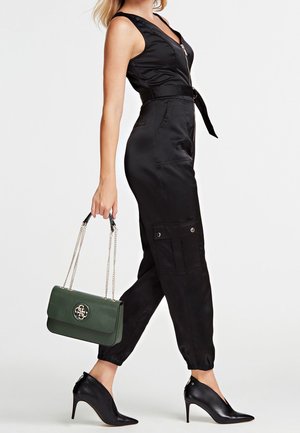 Kobieta ubrana w czarny kombinezon bez rękawów z paskiem, czarne buty na wysokim obcasie, trzyma ciemnozieloną torebkę na łańcuszku.