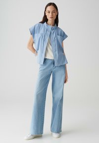 Blauwe korte mouwen jacket met twee zakken, ritsluiting, gedragen over een wit topje en gecombineerd met wijd uitlopende lichtblauwe jeans en witte sneakers.