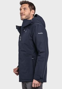 Marineblauwe waterdichte jas met een gestructureerde afwerking, verstelbare capuchon, ritssluiting aan de voorkant en zijzakken. Voorzien van een logo op de mouw.