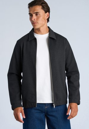 Mand iført en mørkegrå lynlåstrøje over en hvid t-shirt med blå jeans, stående mod en lysegrå baggrund.