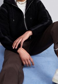 Schwarze Zip-Jacke mit plüschiger Textur und zwei schwarzen Streifen an den Ärmeln, getragen über einem weißen Hemd. Das Modell sitzt auf einer hellblauen Oberfläche.