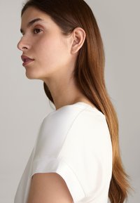 White blouse with a smooth texture, round neckline, and short sleeves, worn by a person with long, straight brown hair. Side profile view.