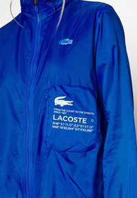 Modrá bunda Lacoste s kapsou na zip na hrudi, zobrazující bílé logo krokodýla a text včetně souřadnic a sloganu "Z kurtu do ulic."