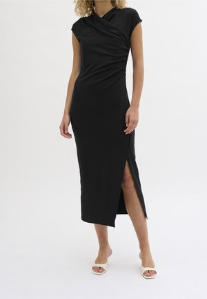 Femme portant une robe midi noire sans manches avec une fente latérale et un détail froncé, associée à des talons ouverts blancs.