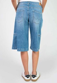 Culottes en denim bleu clair, avec une coupe ample, deux poches arrière avec un design en zigzag, et des ourlets cousus. Portés avec des baskets blanches.