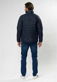 Mørkeblå dunjakke med høy krage og horisontal søm, sammen med blå jeans og hvite sneakers, sett fra baksiden.
