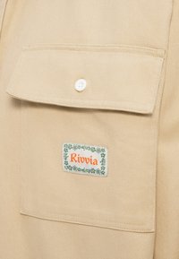 Taschina di tessuto beige con una patta di bottone bianca e un'etichetta rettangolare con la scritta "Rivvia" in arancione, bordata da motivi floreali verdi.