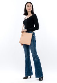 Kvinde iført sort langærmet top og flared jeans, bærende en beige skuldertaske, stående og kiggende til siden på en hvid baggrund.