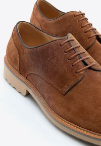 Scarpe in suede marrone con punta arrotondata, lacci e suola in gomma contrastante con finitura testurizzata. Gli accenti includono bordi cuciti.