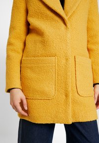 Cappotto giallo con texture, colletto con risvolto, due tasche anteriori e una vestibilità leggermente ampia, abbinato a jeans in denim scuro.