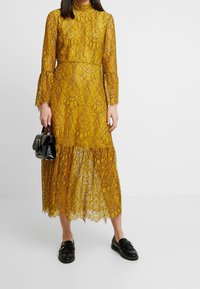 Vestido largo de encaje amarillo con mangas largas, cuello alto, dobladillo con volantes y estampado floral. Combinado con un bolso negro y mocasines negros.