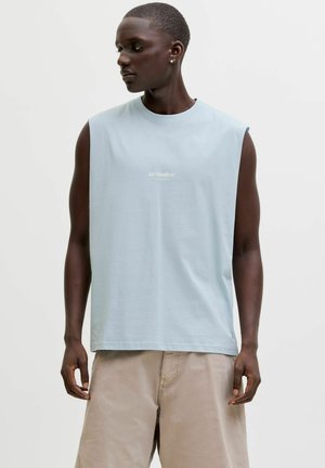 Homme aux cheveux courts portant un débardeur bleu clair avec le texte "JJ Studios" et un short beige, regardant vers sa gauche sur un fond uni.