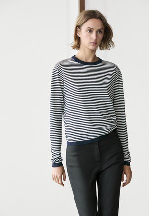 Ung kvinde med kort blondt hår iført en marineblå og hvidstribet langærmet skjorte og sorte bukser, stående mod en neutral baggrund.