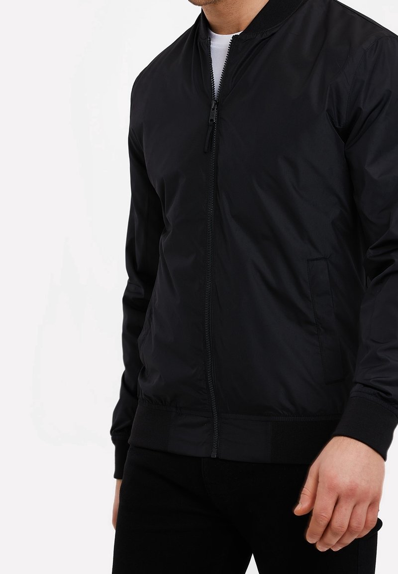 Chaqueta bomber negra con cierre de cremallera frontal, puños y dobladillo acanalados, y bolsillos laterales; hecha de una tela suave y ligera con un acabado mate.