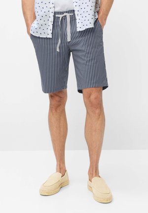 Mand iført navyblå og hvide lodrette stribede shorts med snøre, en hvid skjorte med blå mønstre og beige slip-on sko.